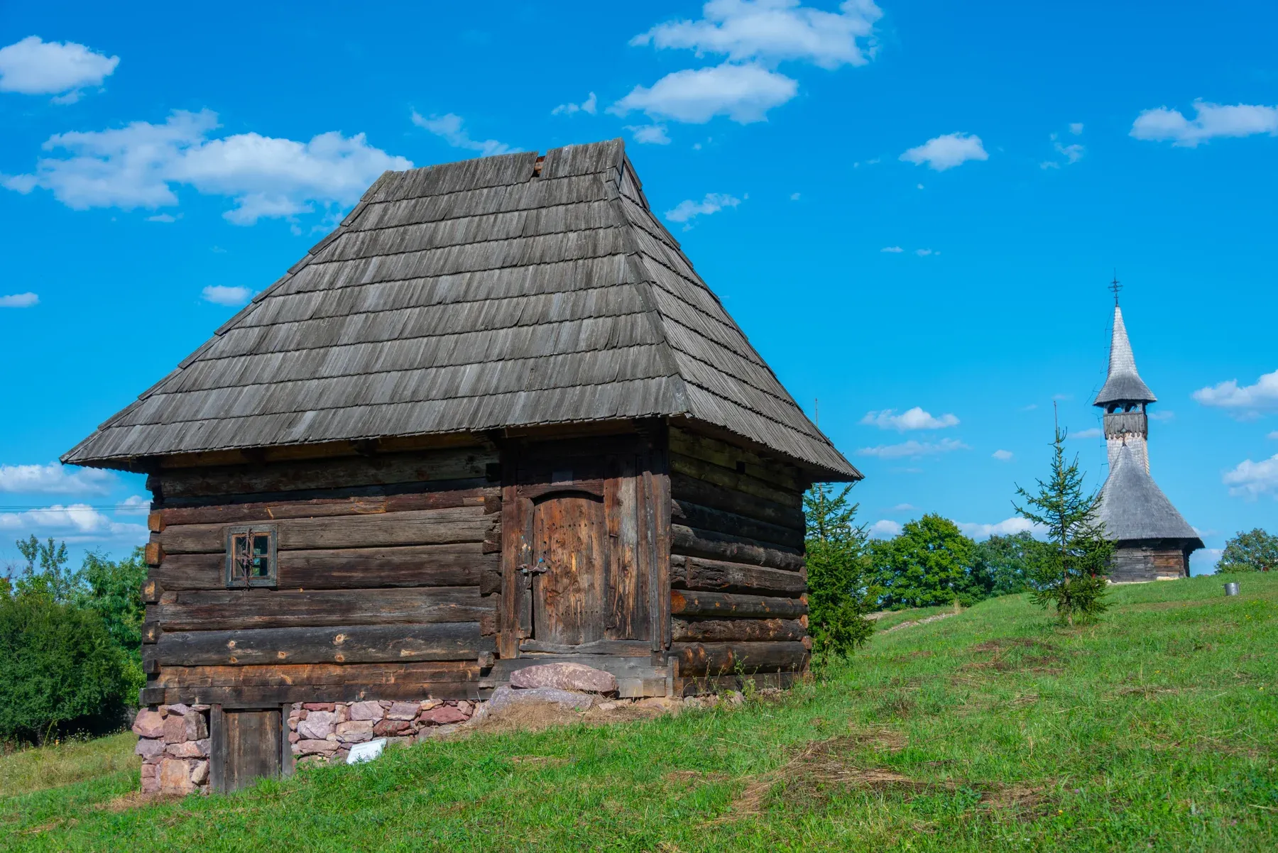 A log house on a stone base; behind it, one of the park's wooden churches